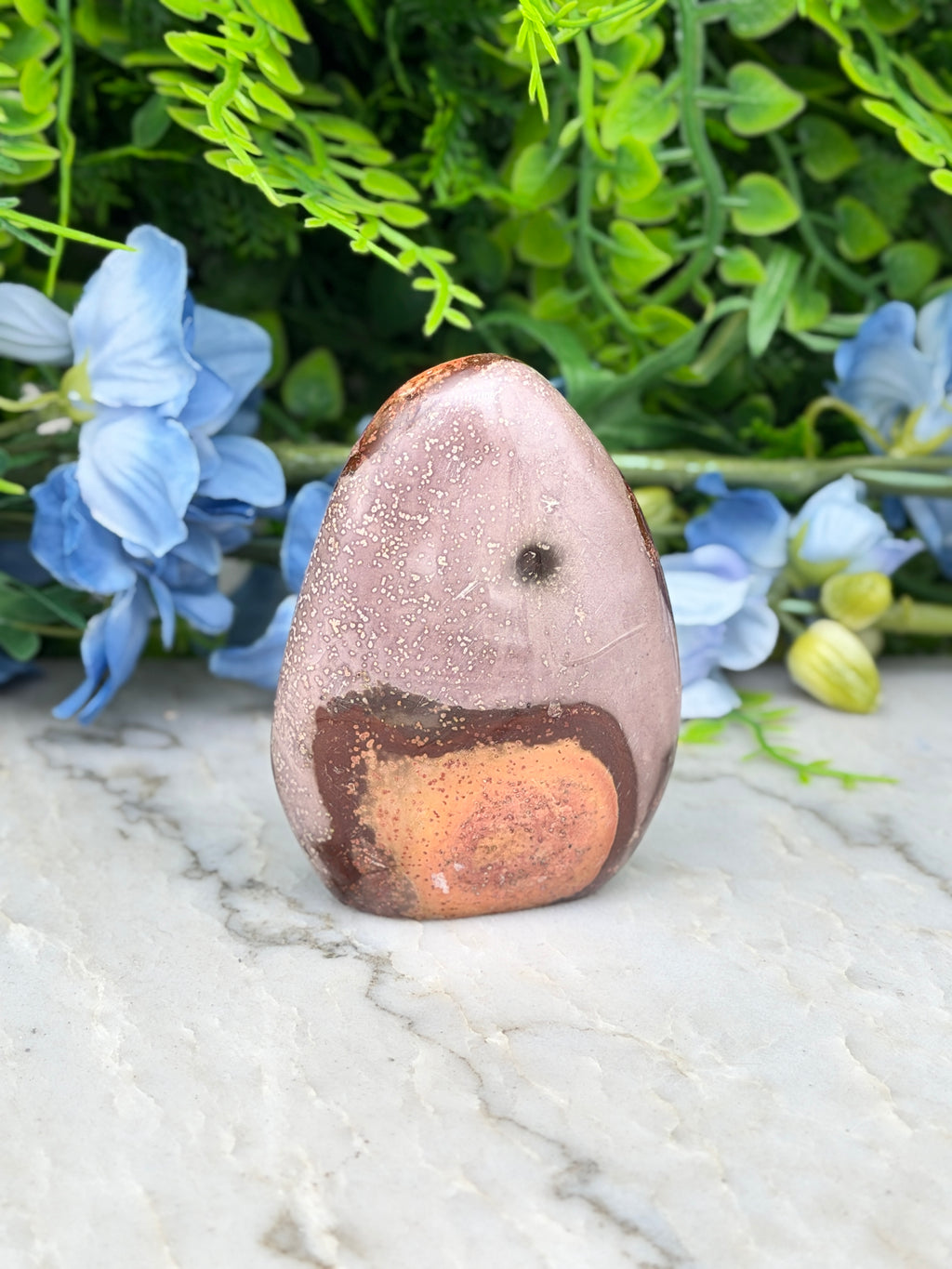 a pinkish-brown rock with a small black spot on it, placed on a white marble surface. The rock is surrounded by blue flowers, creating a visually appealing contrast.