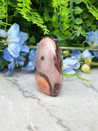 A pinkish-brown jadeite gemstone is displayed on a marble surface, surrounded by blue flowers and greenery.