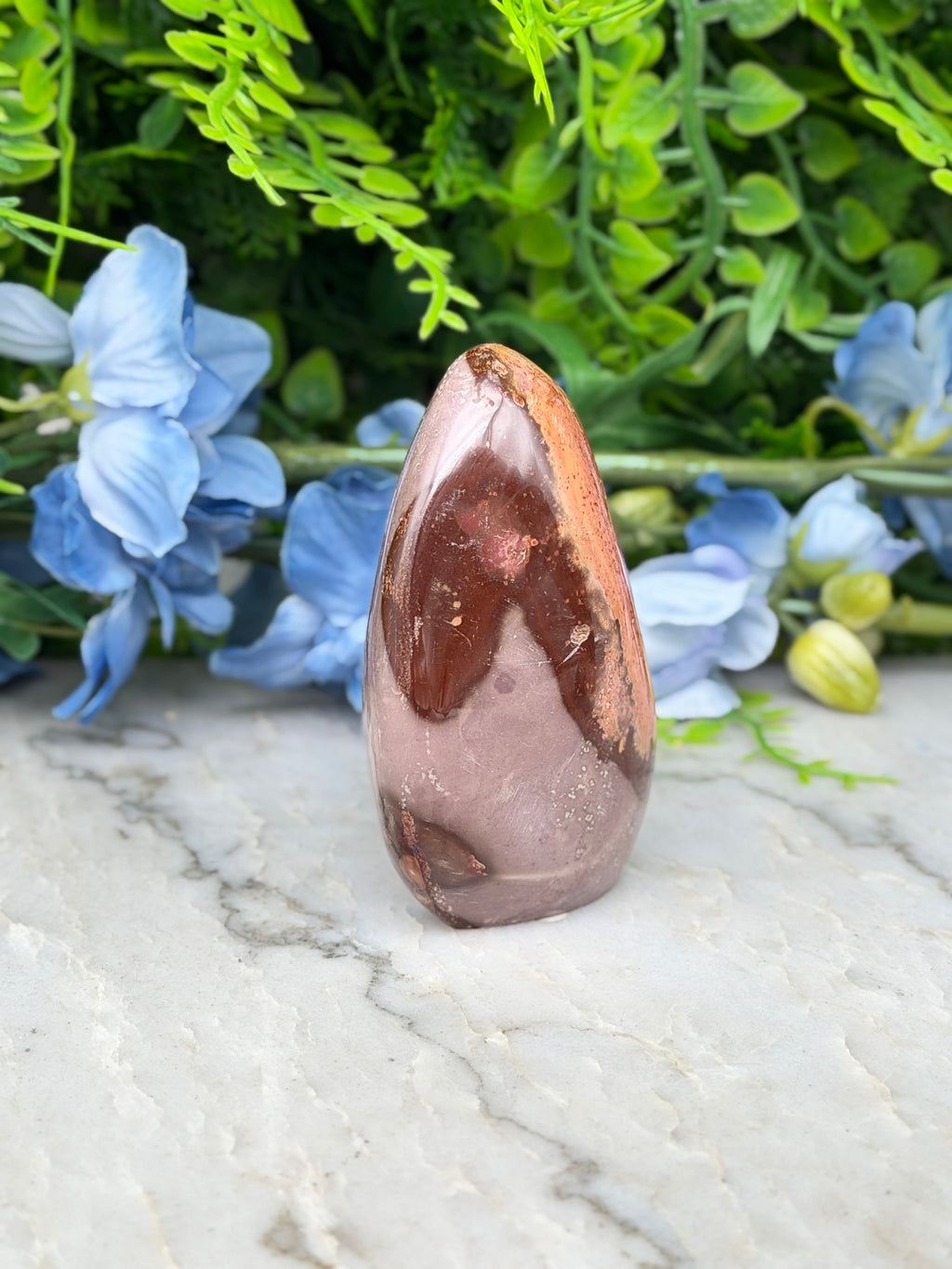 a brown and pink jade crystal surrounded by blue flowers on a marble surface.
