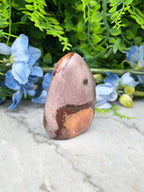 A jadeite stone with a speckled surface sits on a marble surface, surrounded by blue flowers and green foliage.