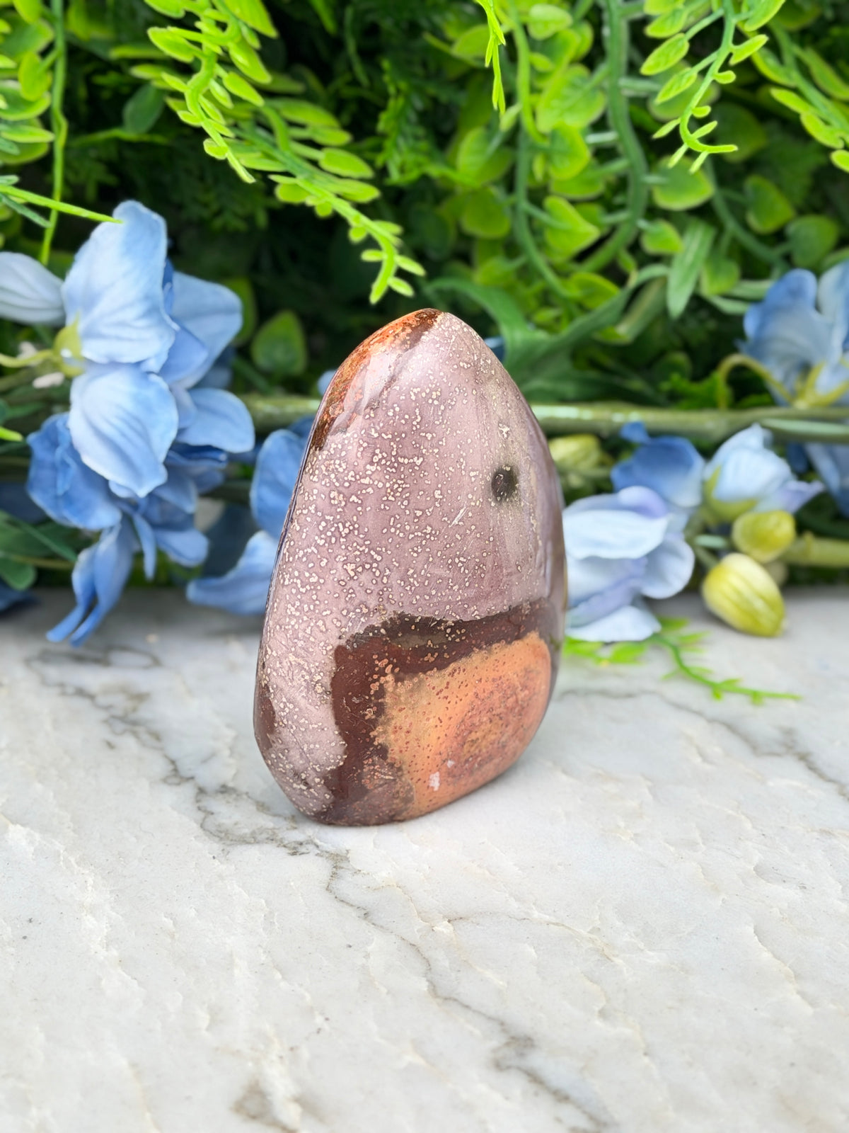 A jadeite stone with a speckled surface sits on a marble surface, surrounded by blue flowers and green foliage.
