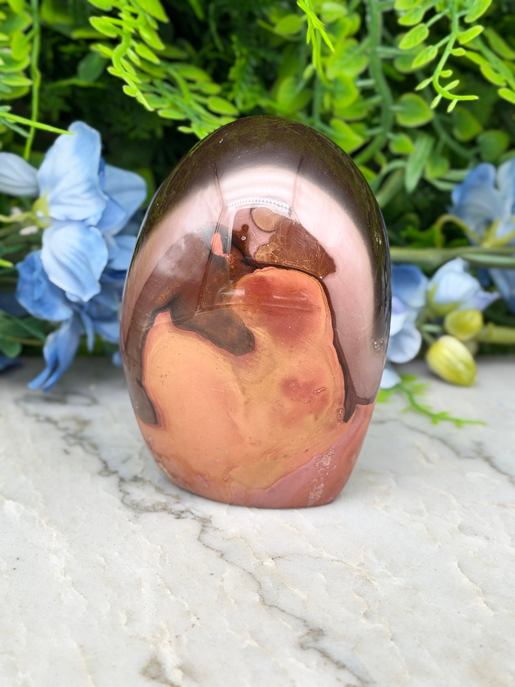 a unique, colorful rock formation with a striking pattern of orange and brown hues, set against a backdrop of lush green foliage and delicate blue flowers.