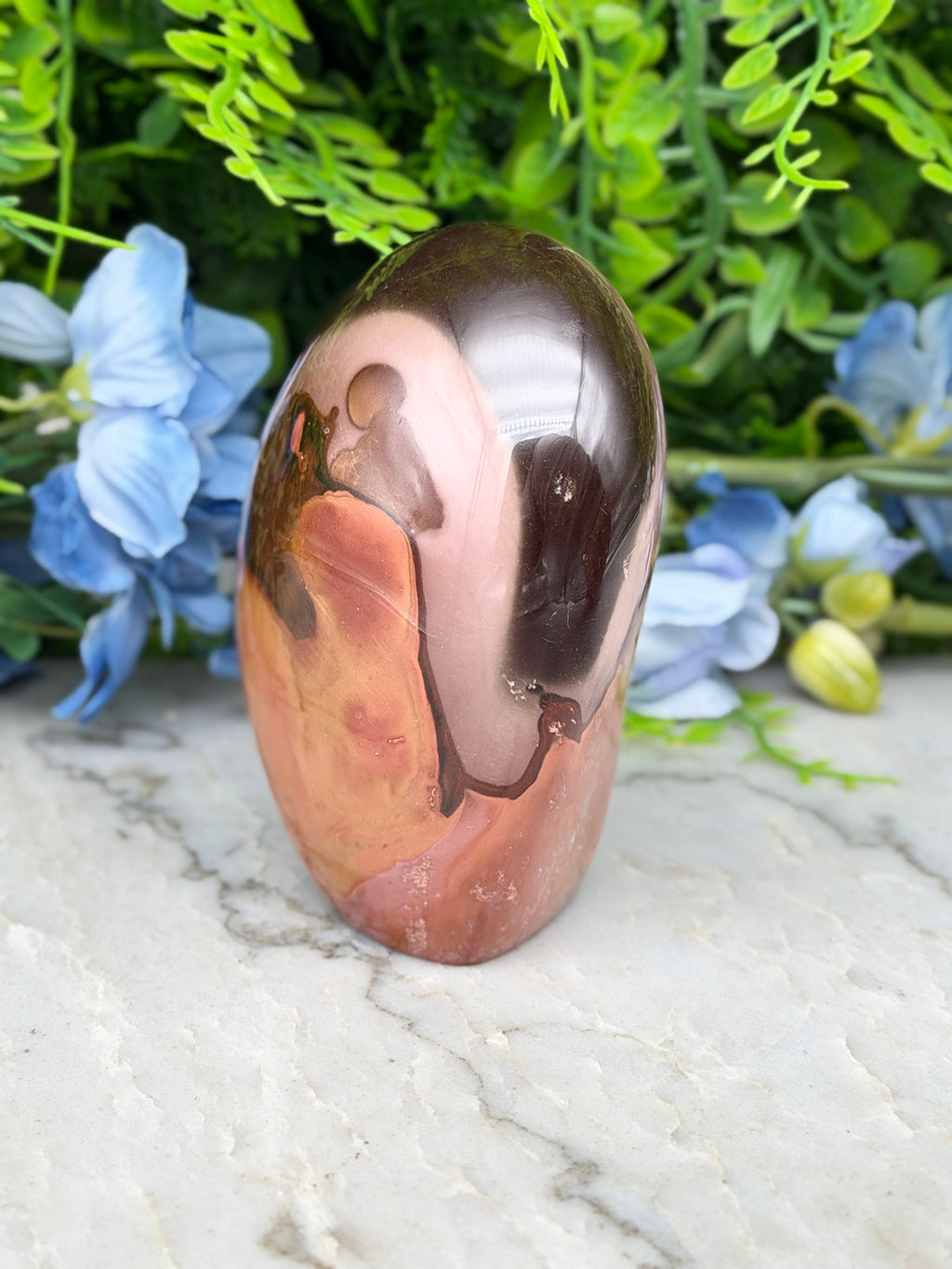 A pinkish-brown jadeite gemstone with a dark brown hue sits on a white marble surface, surrounded by blue flowers.