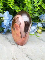 A pinkish-brown jadeite gemstone with a dark brown hue sits on a white marble surface, surrounded by blue flowers.
