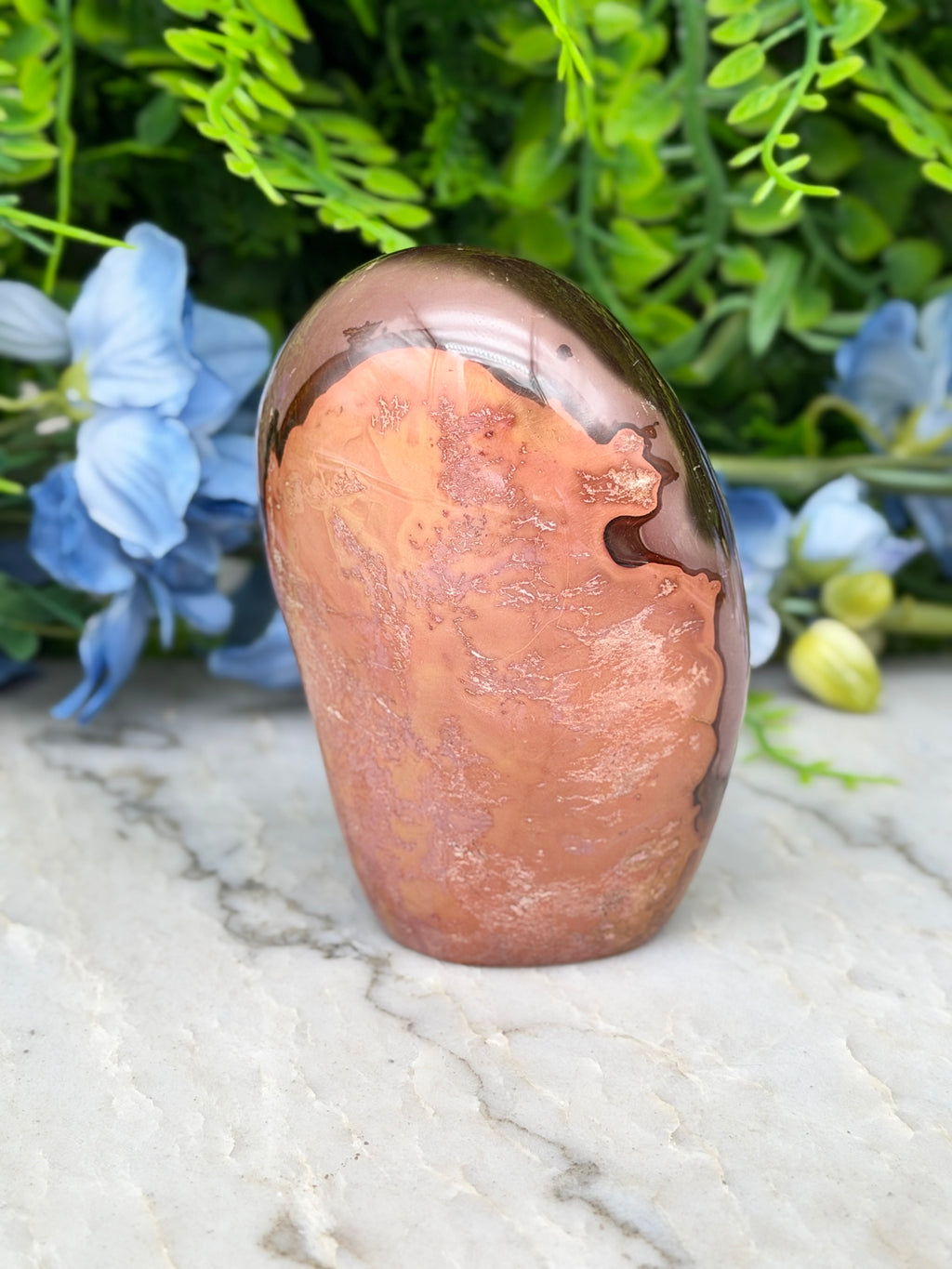 a unique, textured rock formation with a reddish-brown hue, set against a backdrop of lush green foliage and delicate blue flowers.
