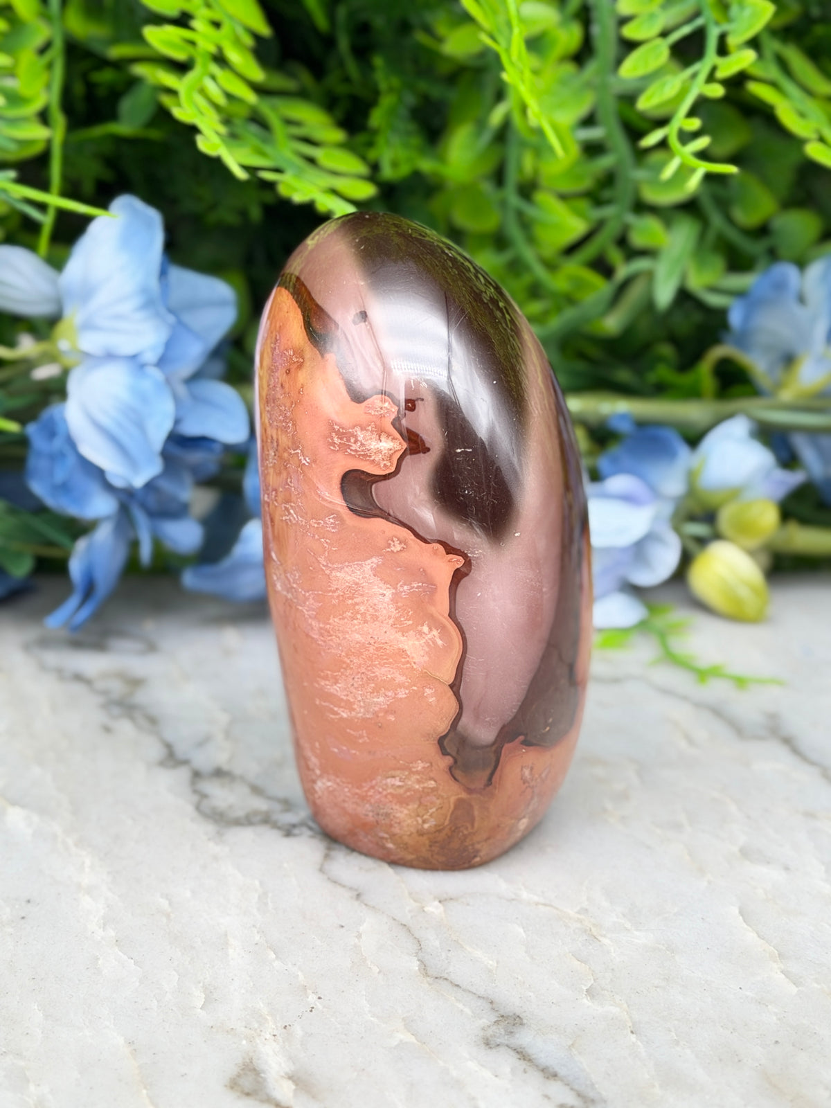 a unique, natural stone with a striking pattern of orange and brown hues, set against a backdrop of lush green foliage and delicate blue flowers.