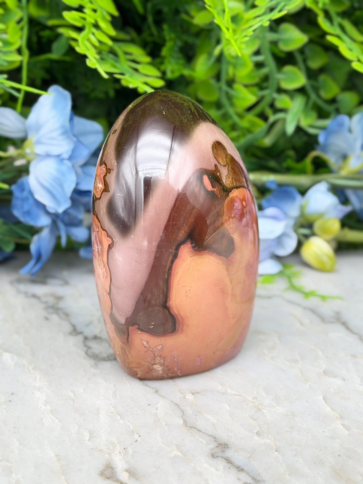A jadeite, a type of jadeite gemstone, is displayed on a marble surface, surrounded by blue flowers.