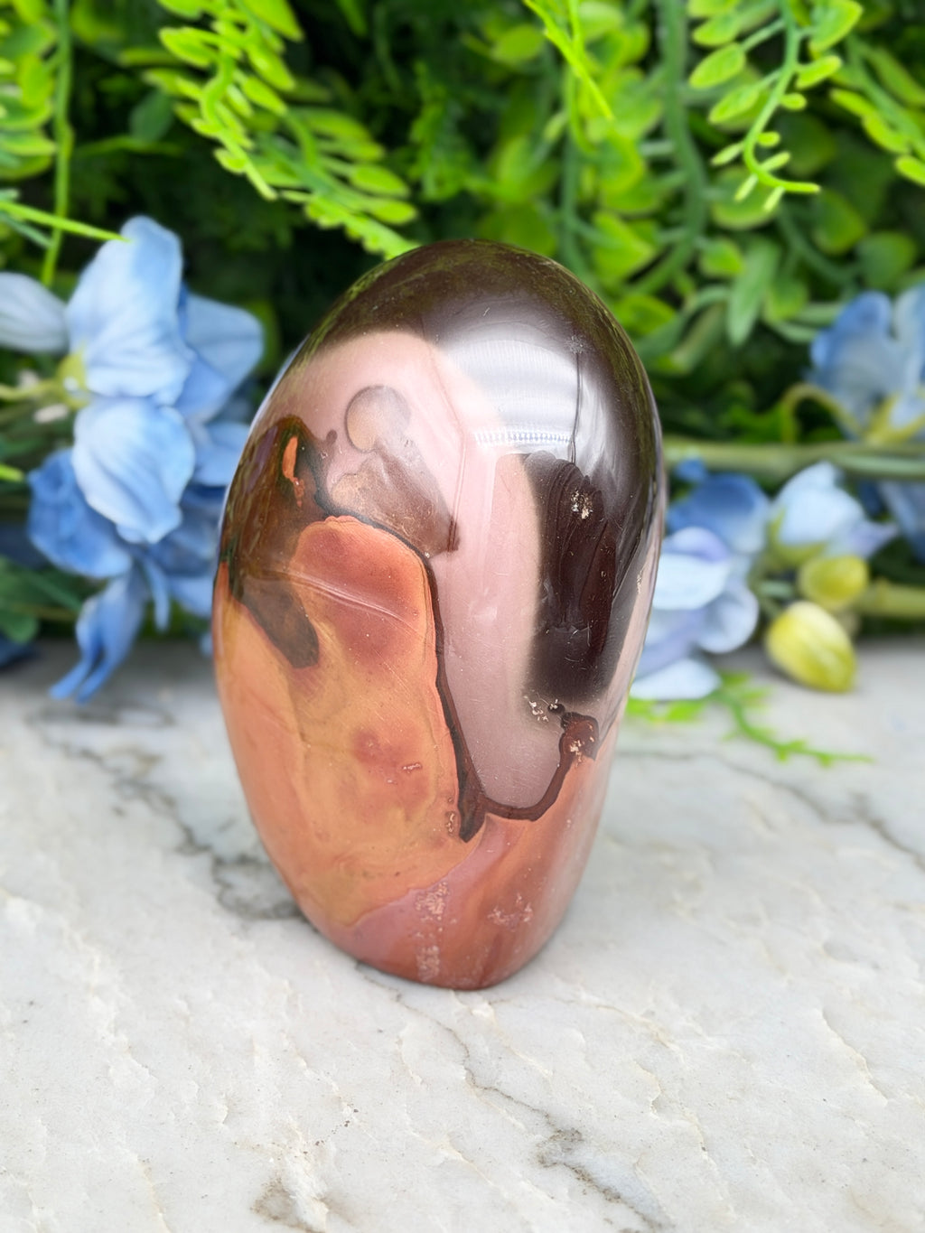 a jadeite, a type of jadeite gemstone, with a unique marbled pattern. The jadeite is placed on a marble surface, surrounded by blue flowers, creating a visually appealing contrast.