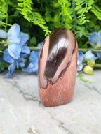 a copper-colored stone or gemstone, which appears to be a piece of jade, placed on a marble surface. The stone has a shiny, reflective surface that catches the light. In the background, there are blue flowers, adding a touch of color and natural beauty to the scene.