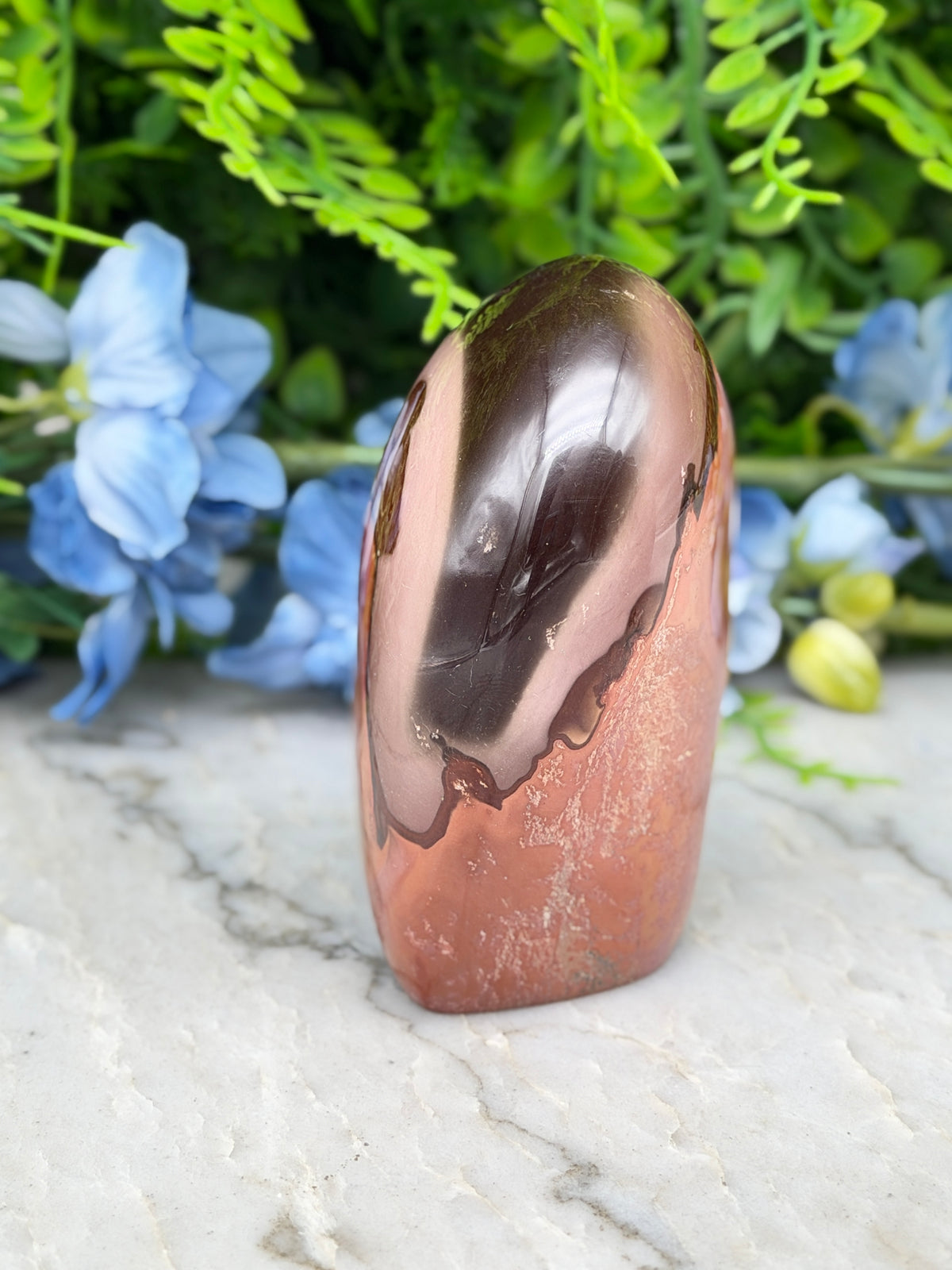 a copper-colored stone or gemstone, which appears to be a piece of jade, placed on a marble surface. The stone has a shiny, reflective surface that catches the light. In the background, there are blue flowers, adding a touch of color and natural beauty to the scene.