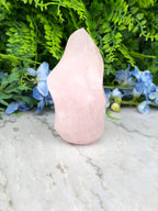 A pink crystal sits on a marble surface, surrounded by lush green foliage and blue flowers.