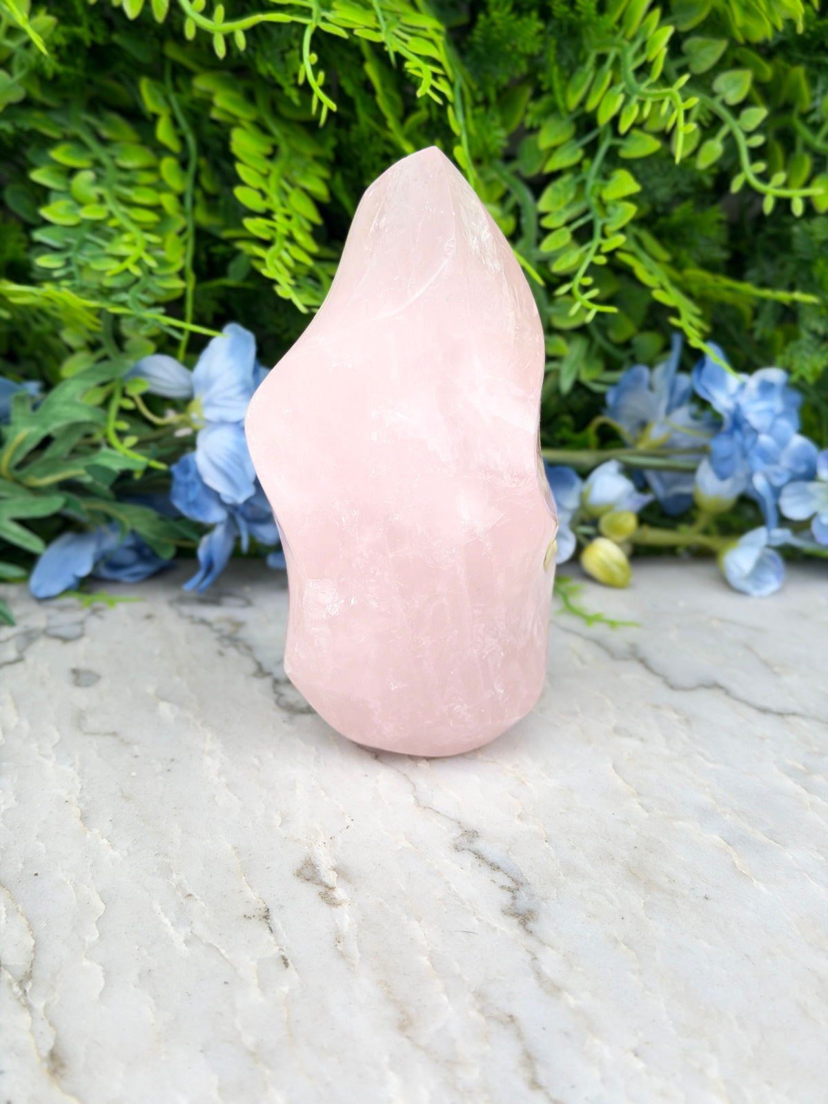 A pink crystal sits on a marble surface, surrounded by lush green foliage and blue flowers.