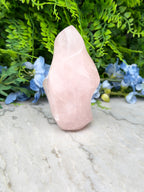 A pink crystal sits on a marble surface, surrounded by blue flowers and lush greenery.