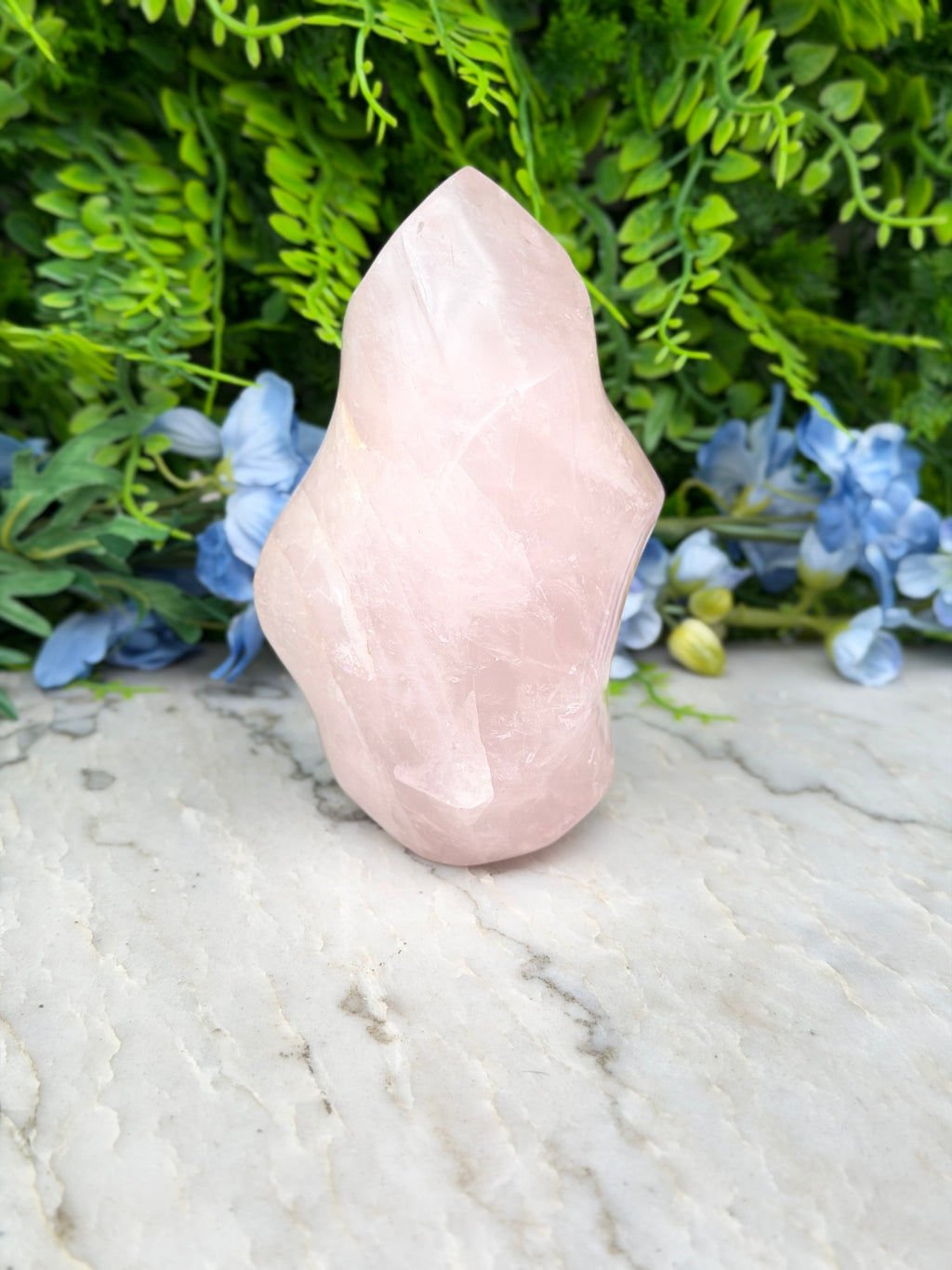 A pink crystal sits on a marble surface, surrounded by lush green foliage and blue flowers.