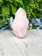 A pink crystal sits on a marble surface, surrounded by lush green foliage and blue flowers.