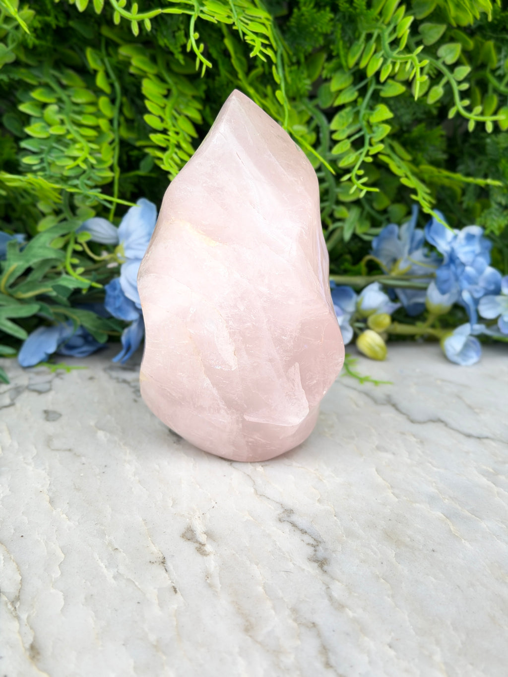 A pink crystal sits on a marble surface, surrounded by lush green foliage and blue flowers.