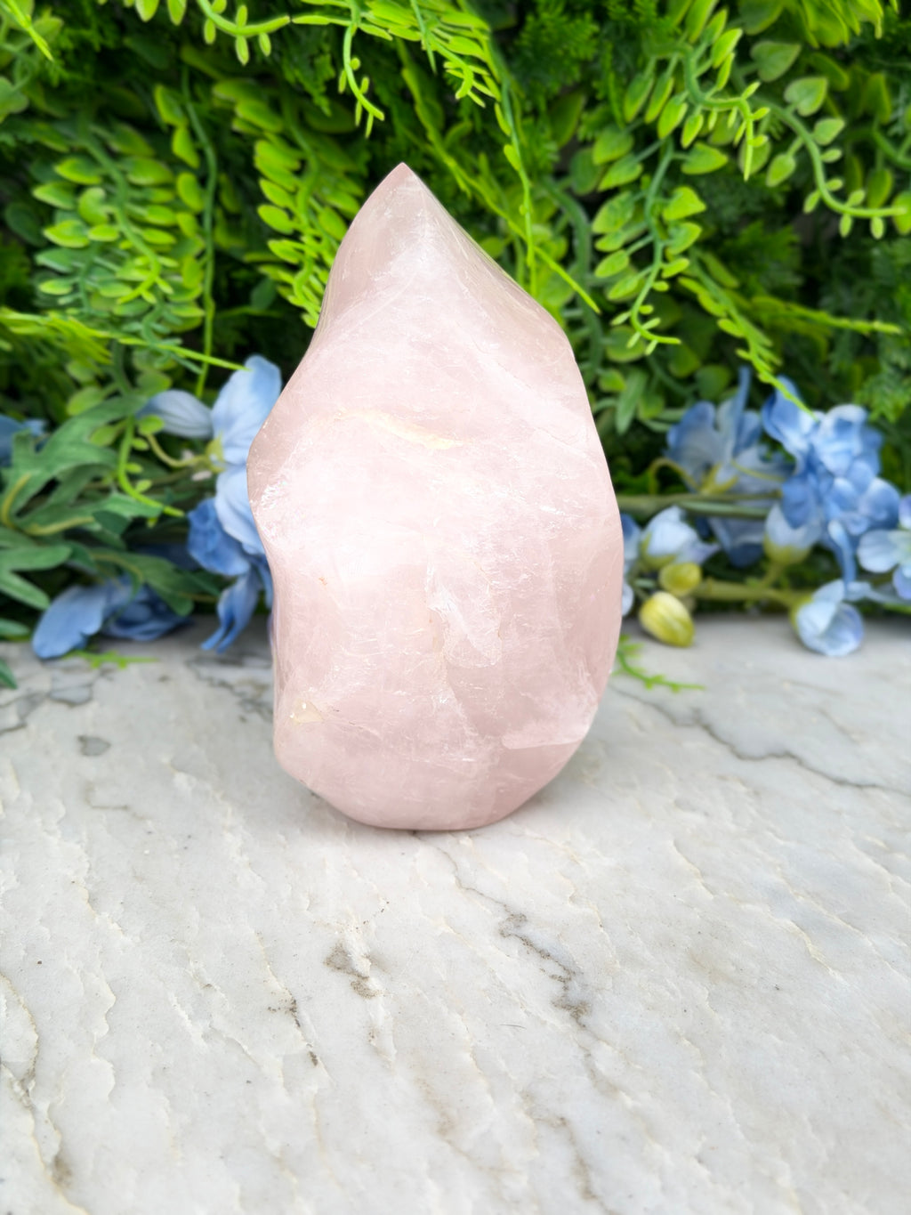 A pink crystal sits on a white marble surface, surrounded by lush green foliage and blue flowers.
