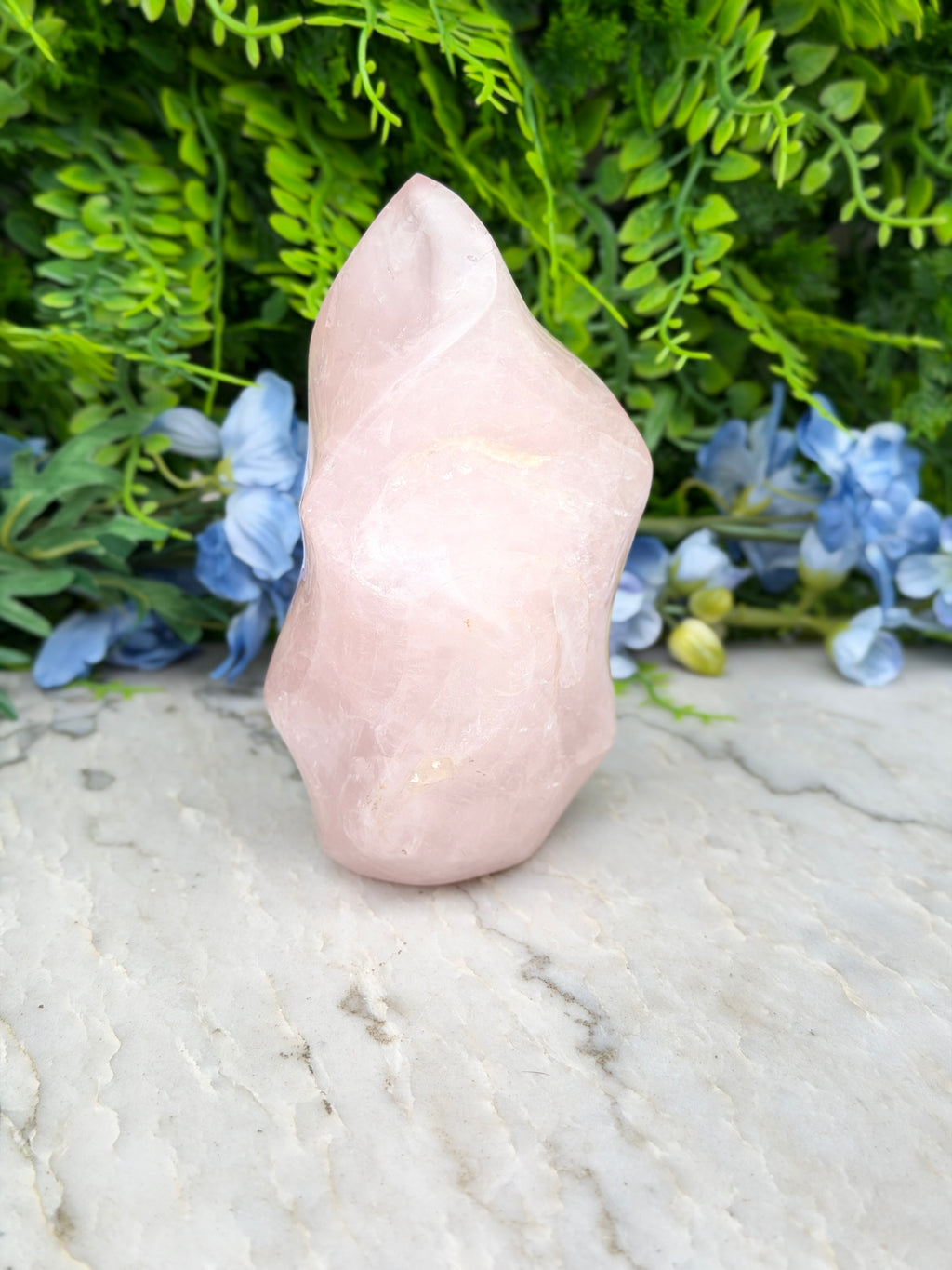 A pink crystal sits on a white marble surface, surrounded by lush green foliage and blue flowers.