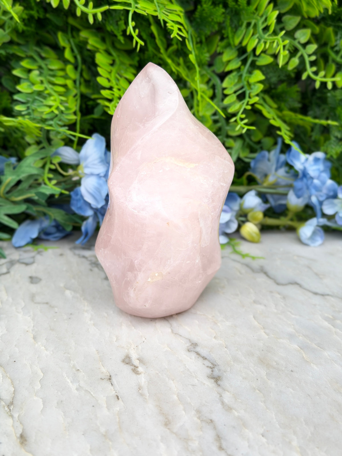 A pink crystal sits on a white marble surface, surrounded by lush green foliage and blue flowers.