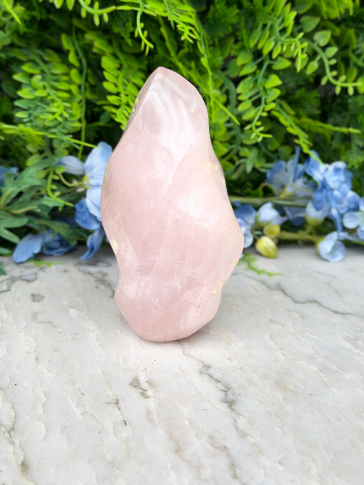A pink crystal sits on a marble surface, surrounded by lush green foliage and blue flowers.