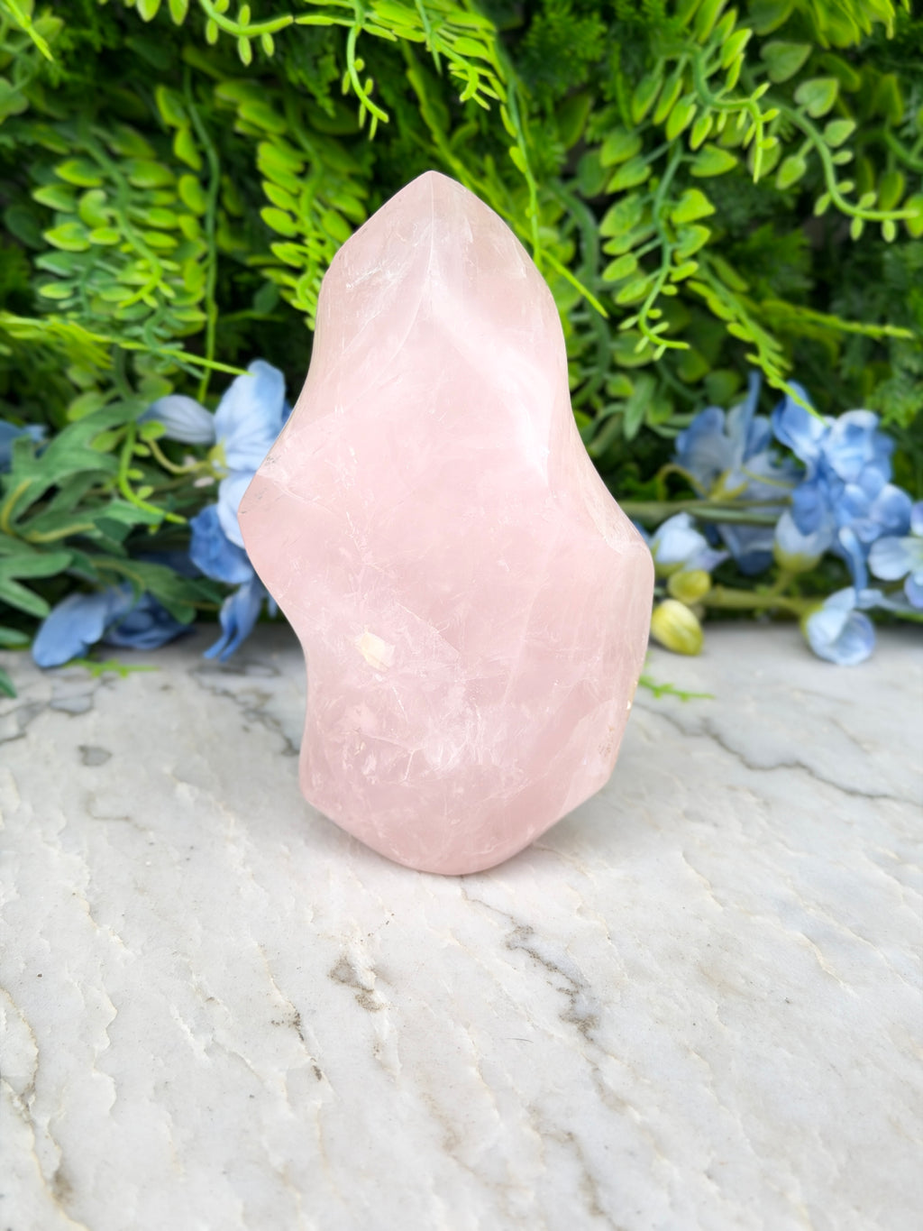 A pink crystal sits on a marble surface, surrounded by lush green foliage and blue flowers.