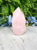 A pink crystal sits on a marble surface, surrounded by lush green foliage and blue flowers.