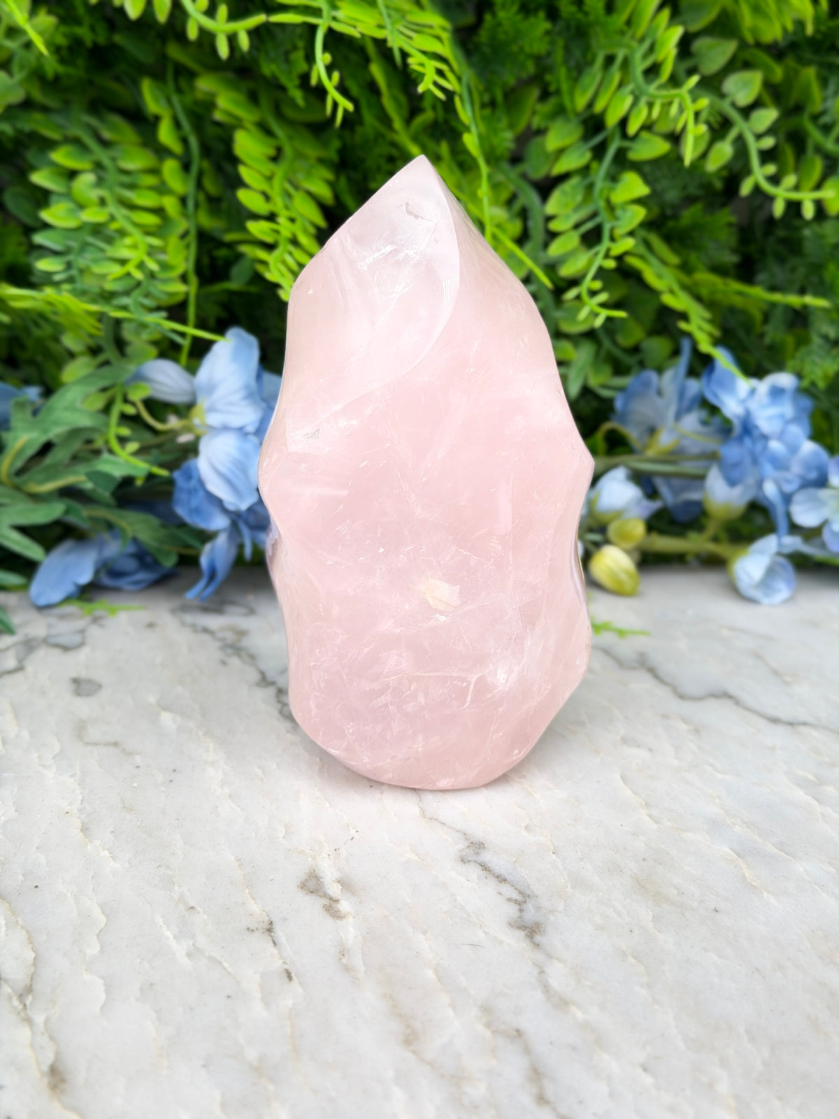 A pink crystal sits on a marble surface, surrounded by lush green foliage and blue flowers.
