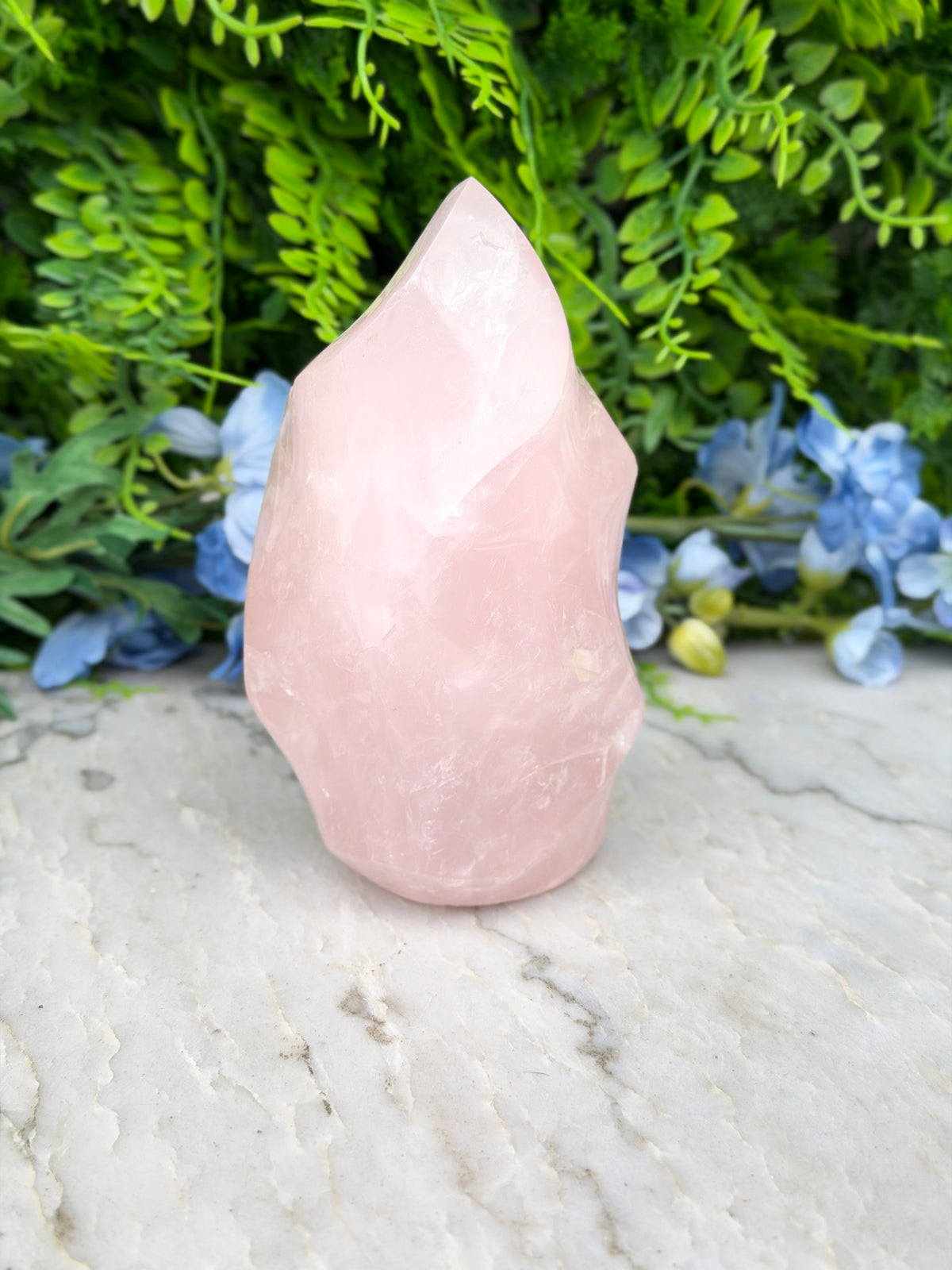 A pink crystal sits on a marble surface, surrounded by lush green foliage and blue flowers.