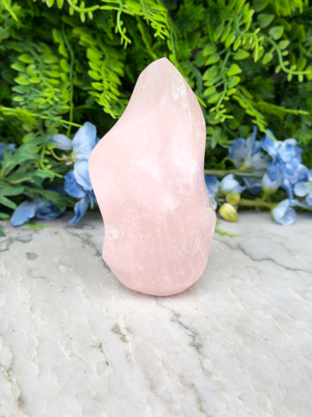 A pink crystal sits on a marble surface, surrounded by lush green foliage and blue flowers.