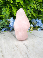 A pink crystal sits on a marble surface, surrounded by blue flowers and lush greenery.