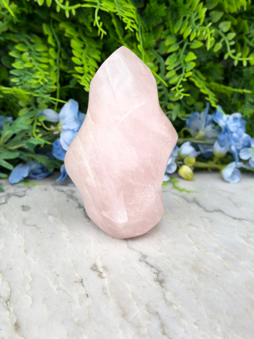 A pink crystal sits on a marble surface, surrounded by lush green foliage and blue flowers.