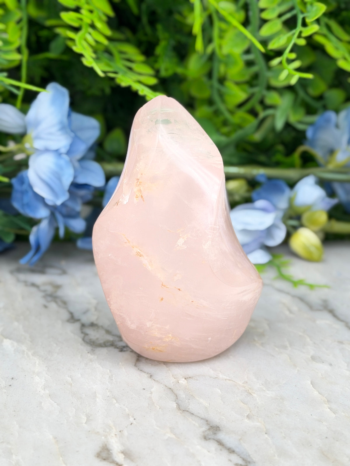 A pink crystal sits on a marble surface, surrounded by blue flowers and green foliage.