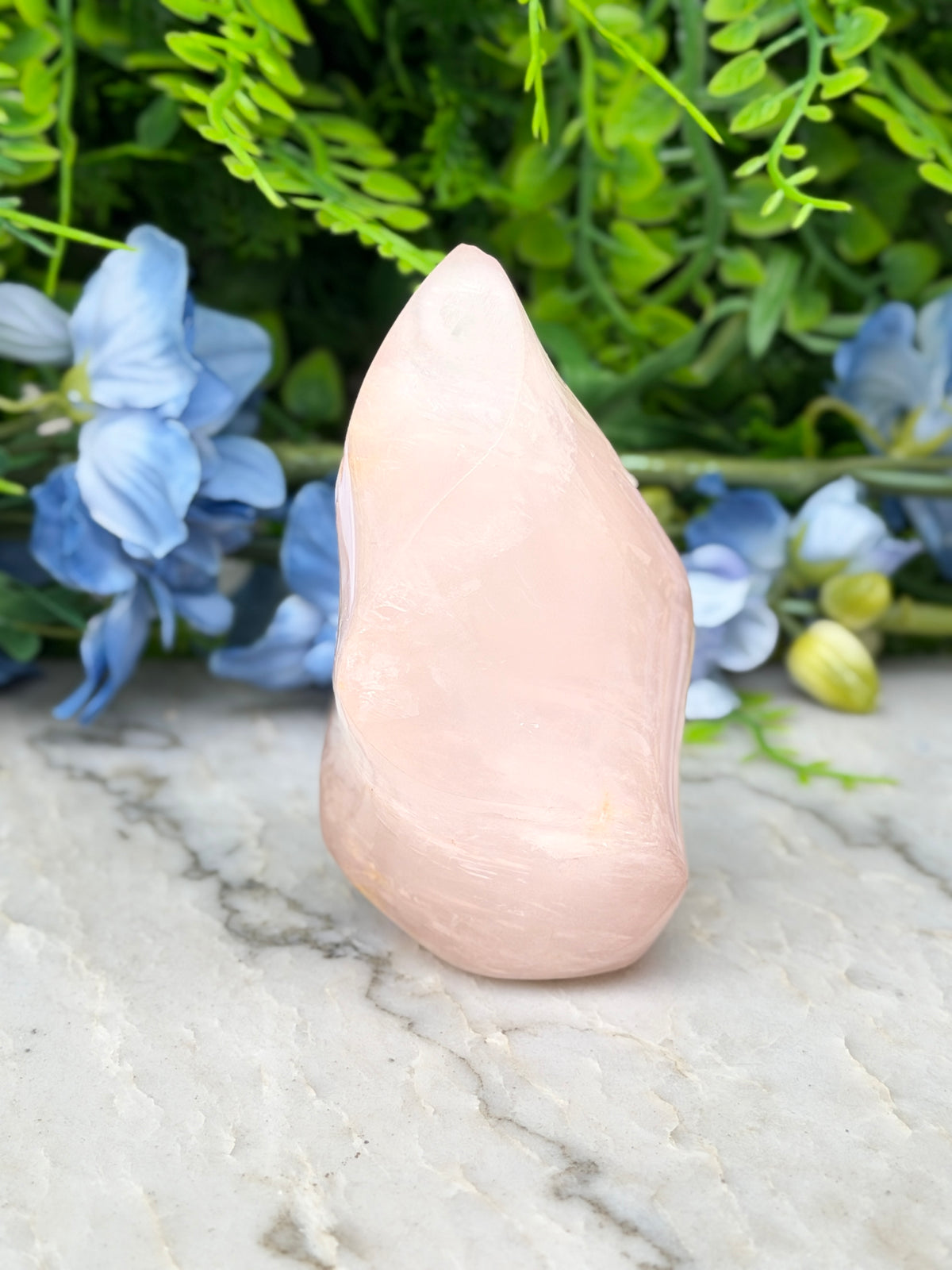A pink crystal sits on a marble surface, surrounded by blue flowers and green foliage.