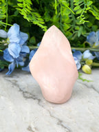 A pink crystal sits on a marble surface, surrounded by blue flowers and green foliage.