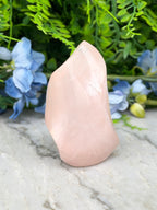 A pink crystal sits on a marble surface, surrounded by blue flowers and green foliage.