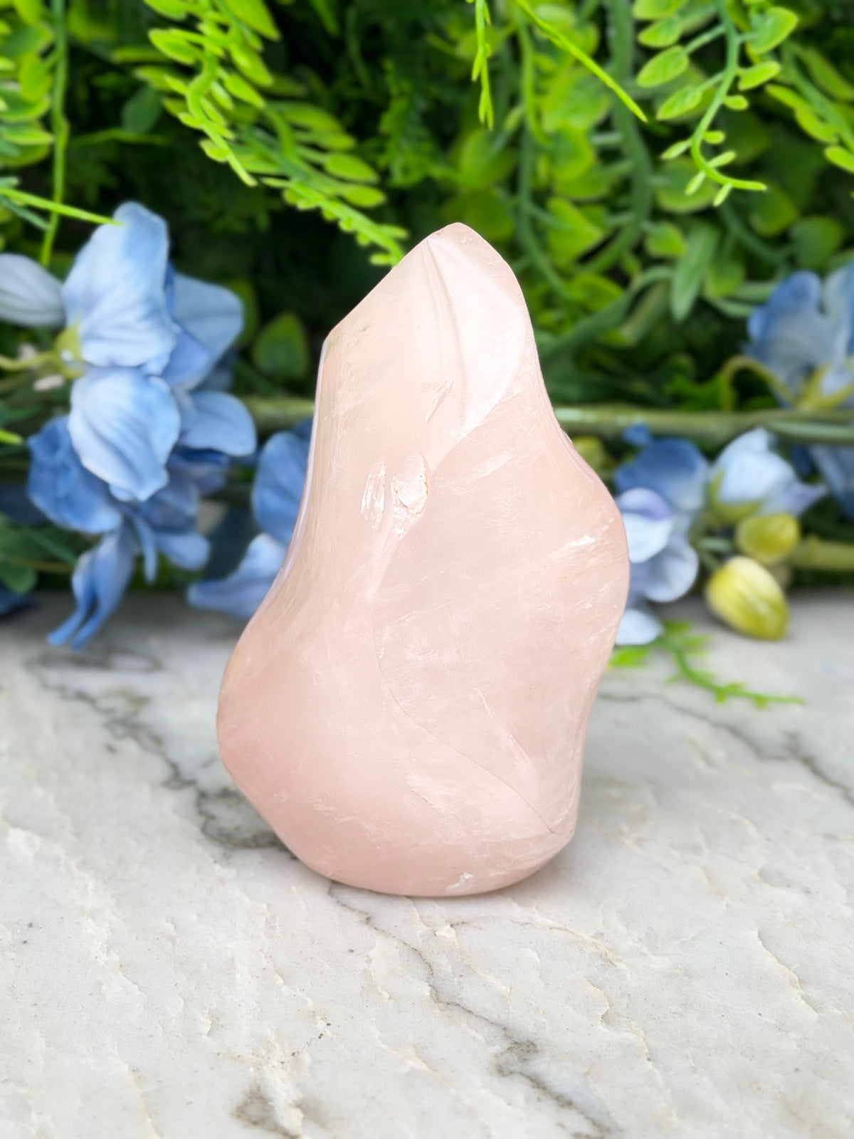 A pink crystal sits on a marble surface, surrounded by blue flowers and green foliage.