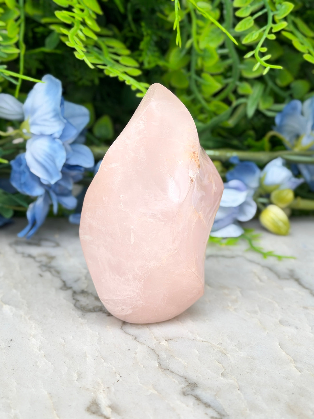 A pink crystal sits on a marble surface, surrounded by blue flowers and green foliage.