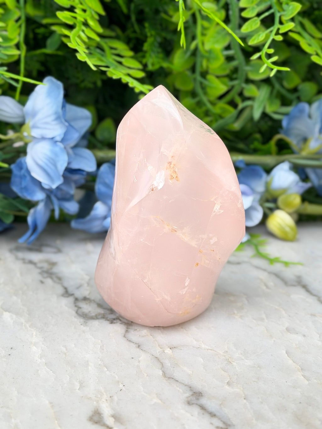 A pink crystal sits on a marble surface, surrounded by blue flowers and green foliage.