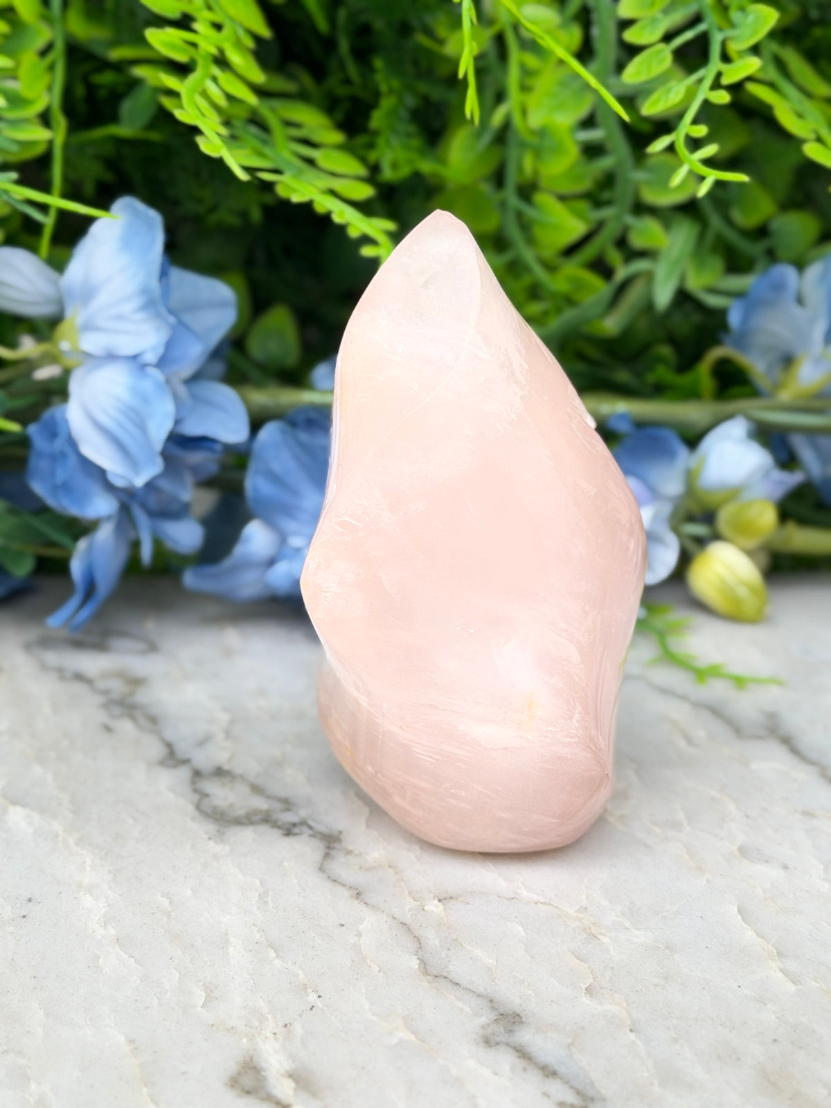 A pink crystal sits on a marble surface, surrounded by blue flowers and green foliage.