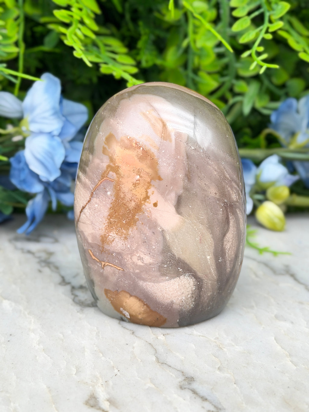 a jadeite, a type of jadeite gemstone, with a unique, colorful, and textured surface. The jadeite is set against a backdrop of lush green foliage, creating a visually striking contrast.
