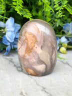 a jadeite, a type of jadeite gemstone, with a unique, colorful, and textured surface. The jadeite is set against a backdrop of lush green foliage, creating a visually striking contrast.