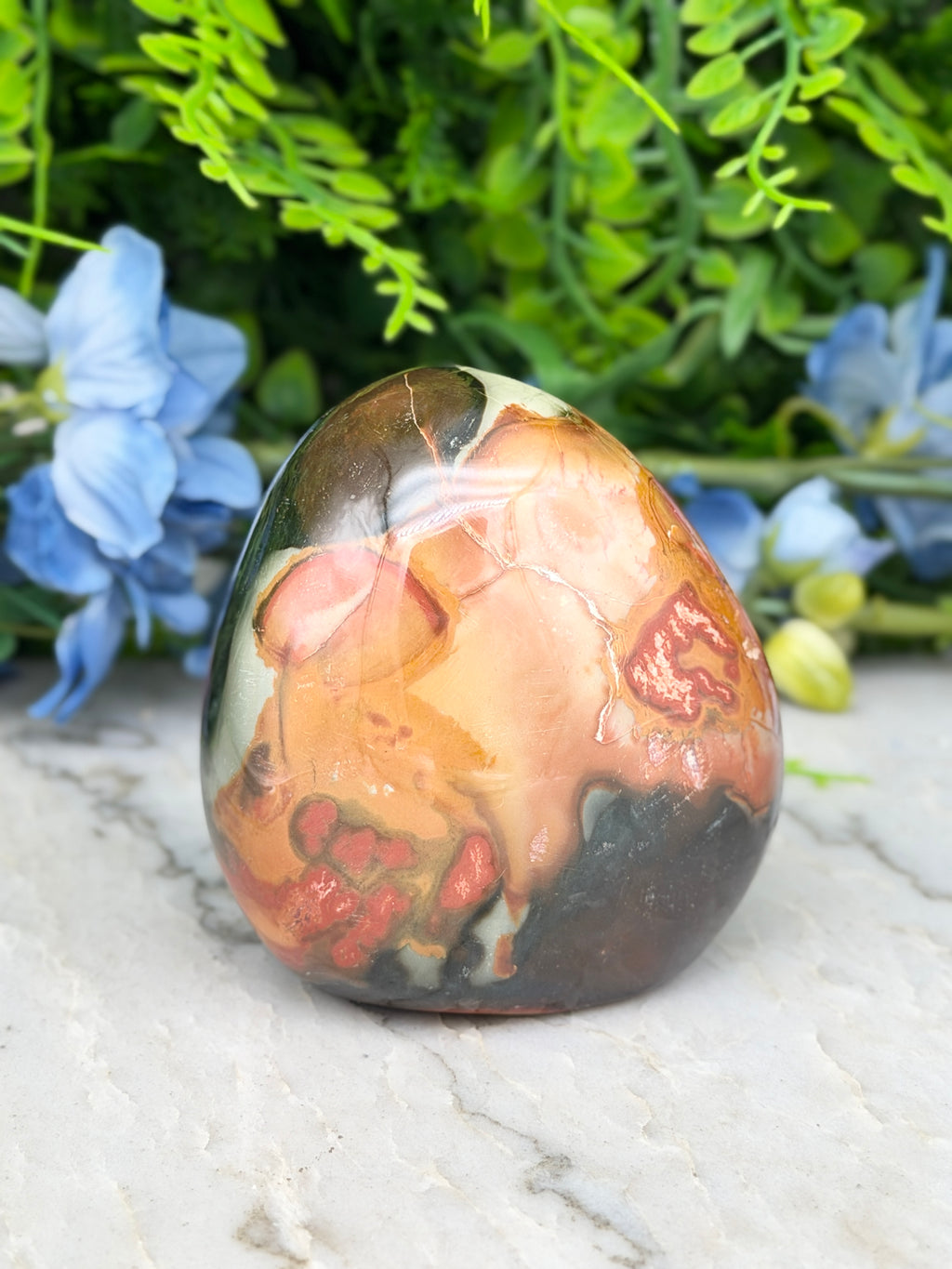 A colorful, abstract-patterned rock sits on a white surface, surrounded by blue flowers and greenery.