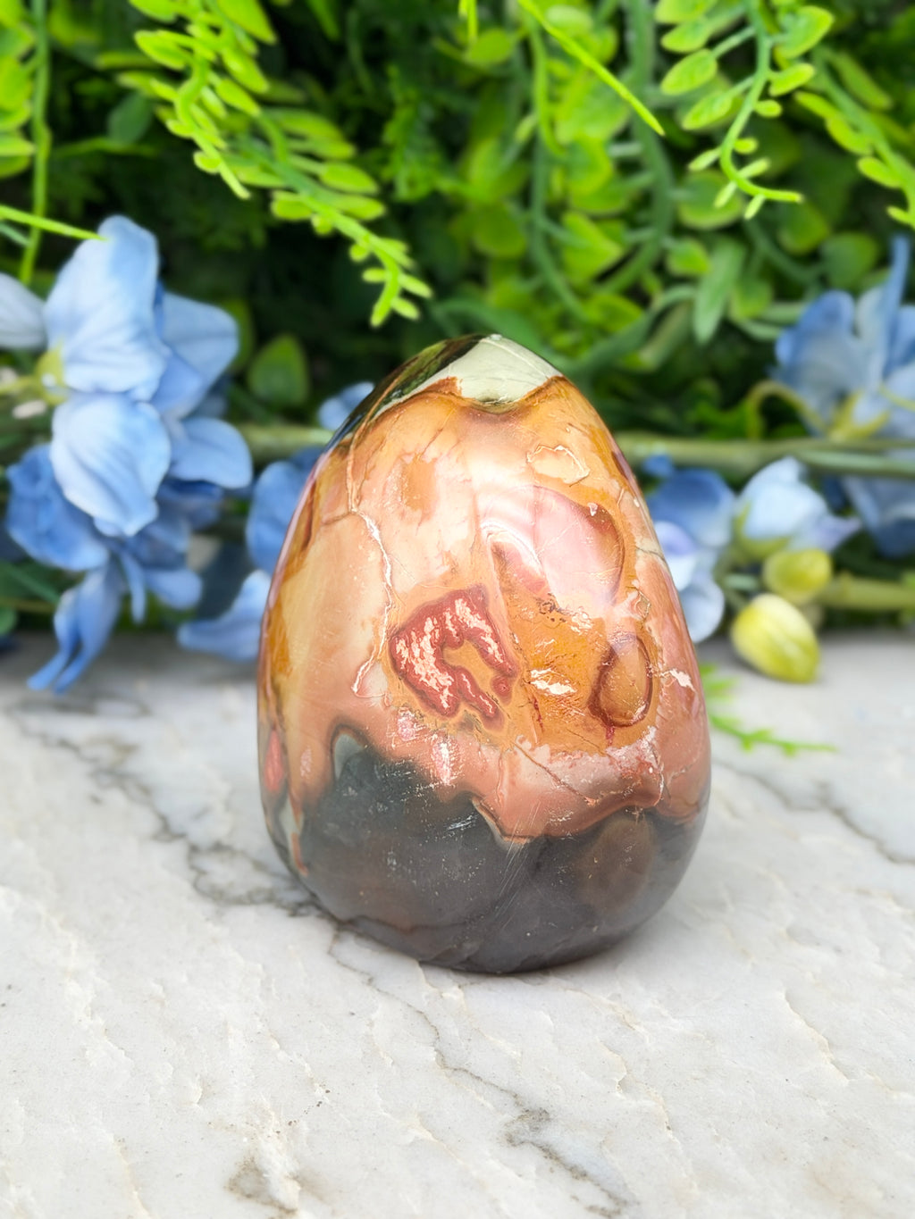 a jadeite, a type of jadeite gemstone, with a unique speckled pattern. The jadeite is placed on a marble surface, surrounded by blue flowers, creating a visually appealing and harmonious composition.