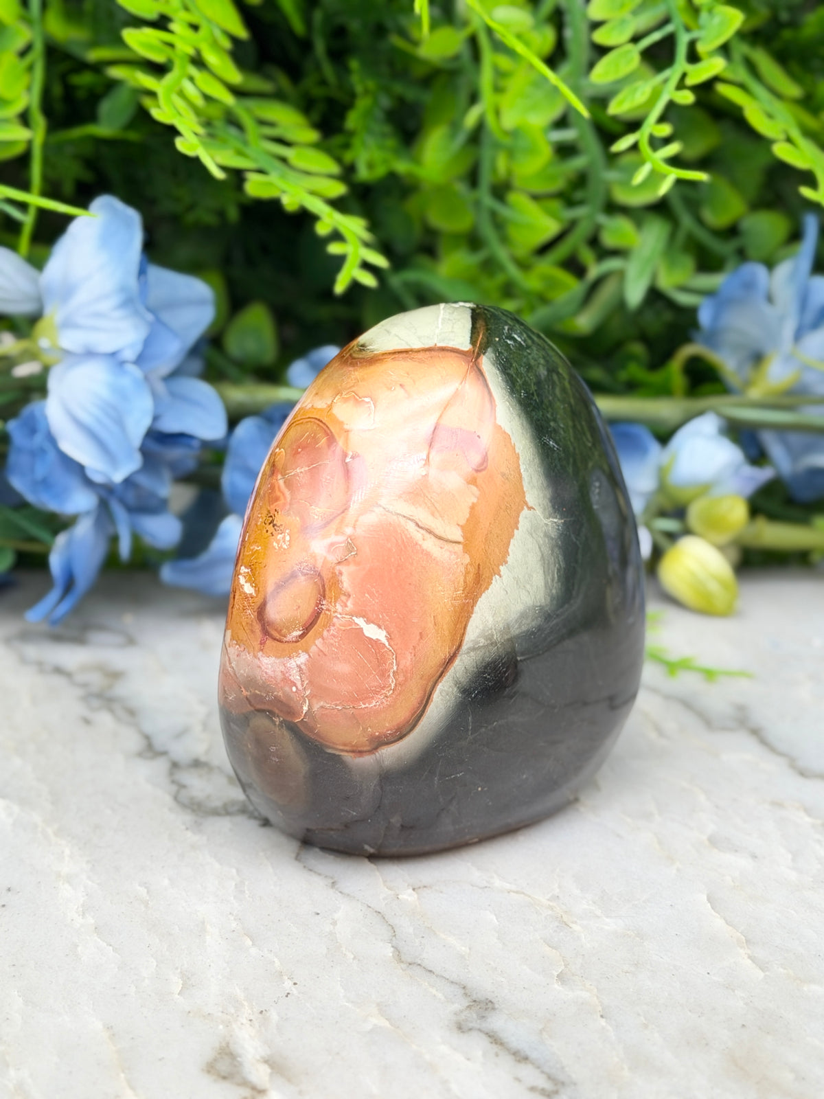 a jade egg, a natural stone with a unique, oval shape, resting on a marble surface. The jade egg has a black and orange hue, with a white and orange pattern on its surface. The background is adorned with blue flowers, adding a touch of color and life to the scene.