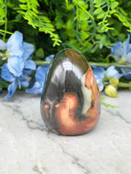 a colorful, textured rock formation with a mix of orange, black, and brown hues, set against a backdrop of lush green foliage and delicate blue flowers.
