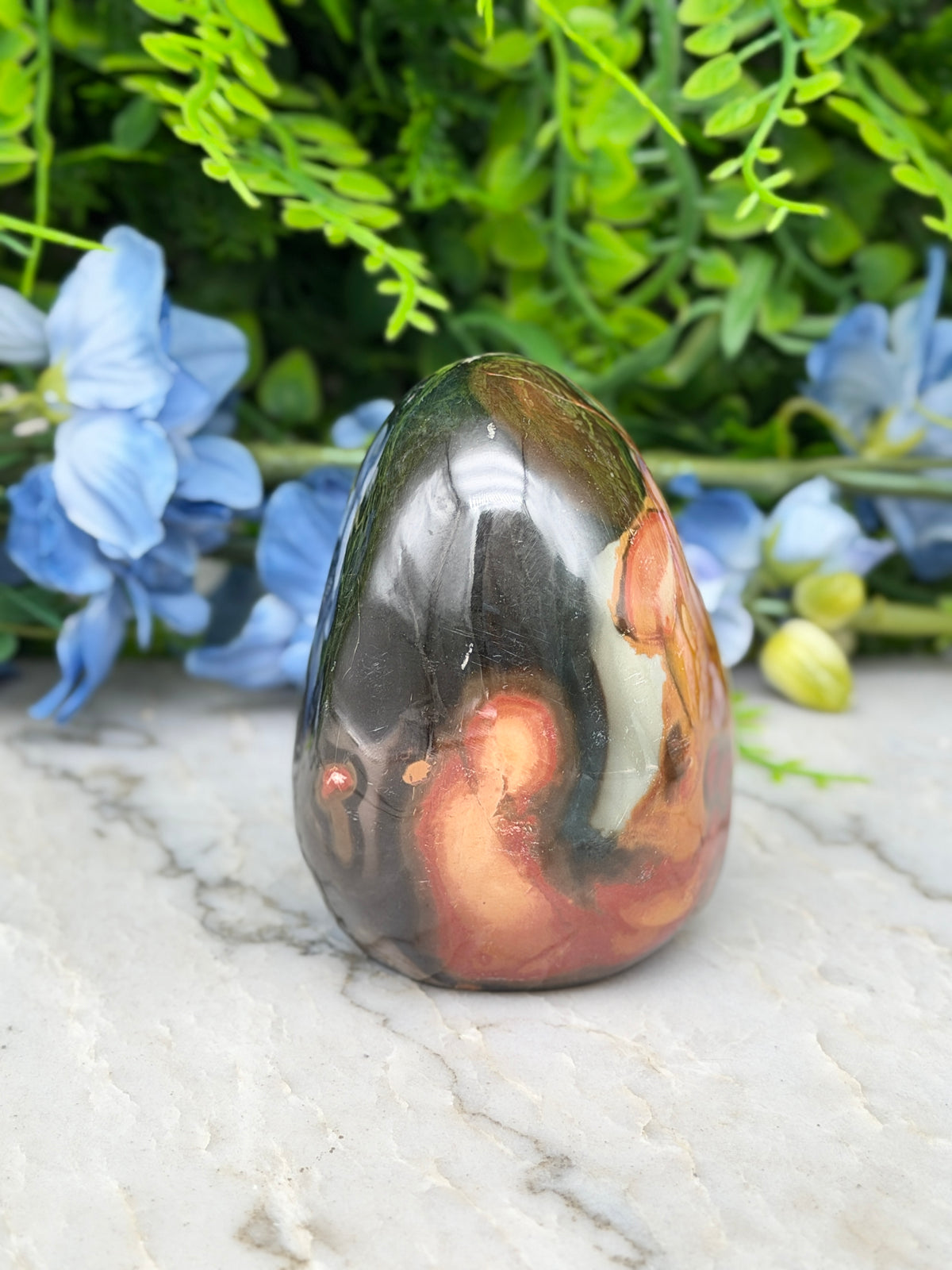 a colorful, textured rock formation with a mix of orange, black, and brown hues, set against a backdrop of lush green foliage and delicate blue flowers.