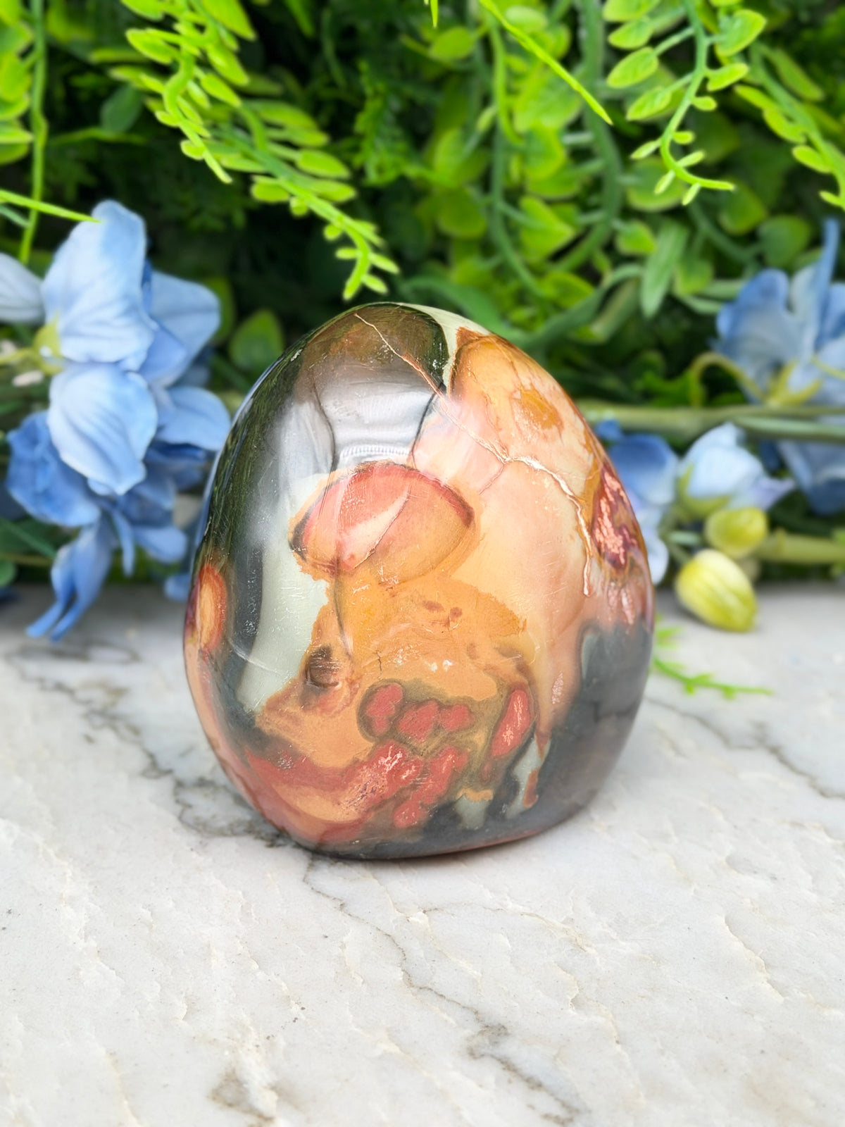 A colorful, abstract-patterned stone or gemstone is displayed on a white surface, surrounded by blue flowers and greenery.