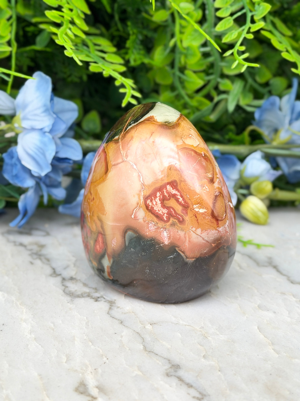 a colorful, textured rock formation with a mix of orange, brown, and black hues, set against a backdrop of lush green foliage and delicate blue flowers.