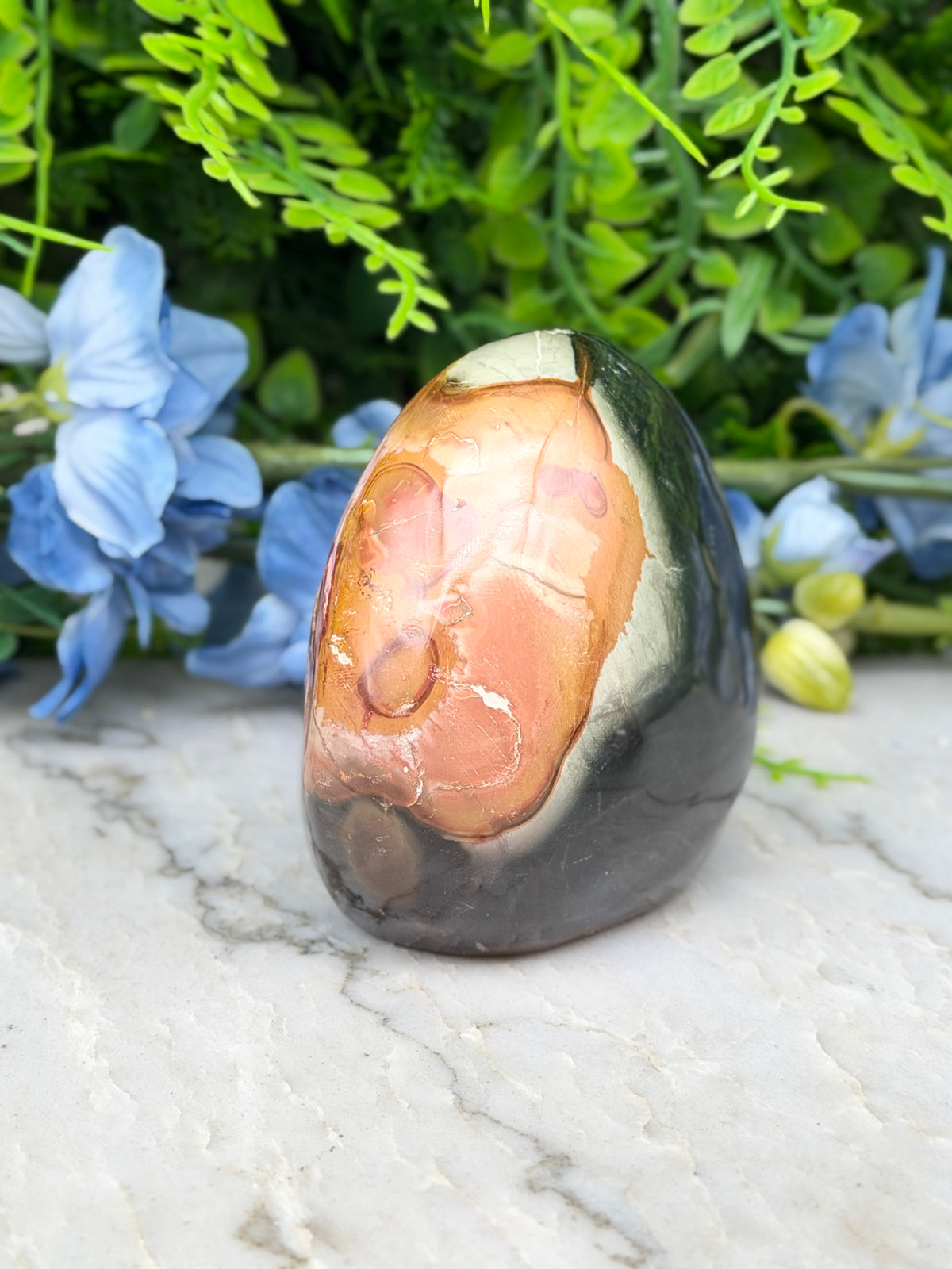A jadeite gemstone with a unique pattern of orange and black hues sits on a white marble surface, surrounded by blue flowers.
