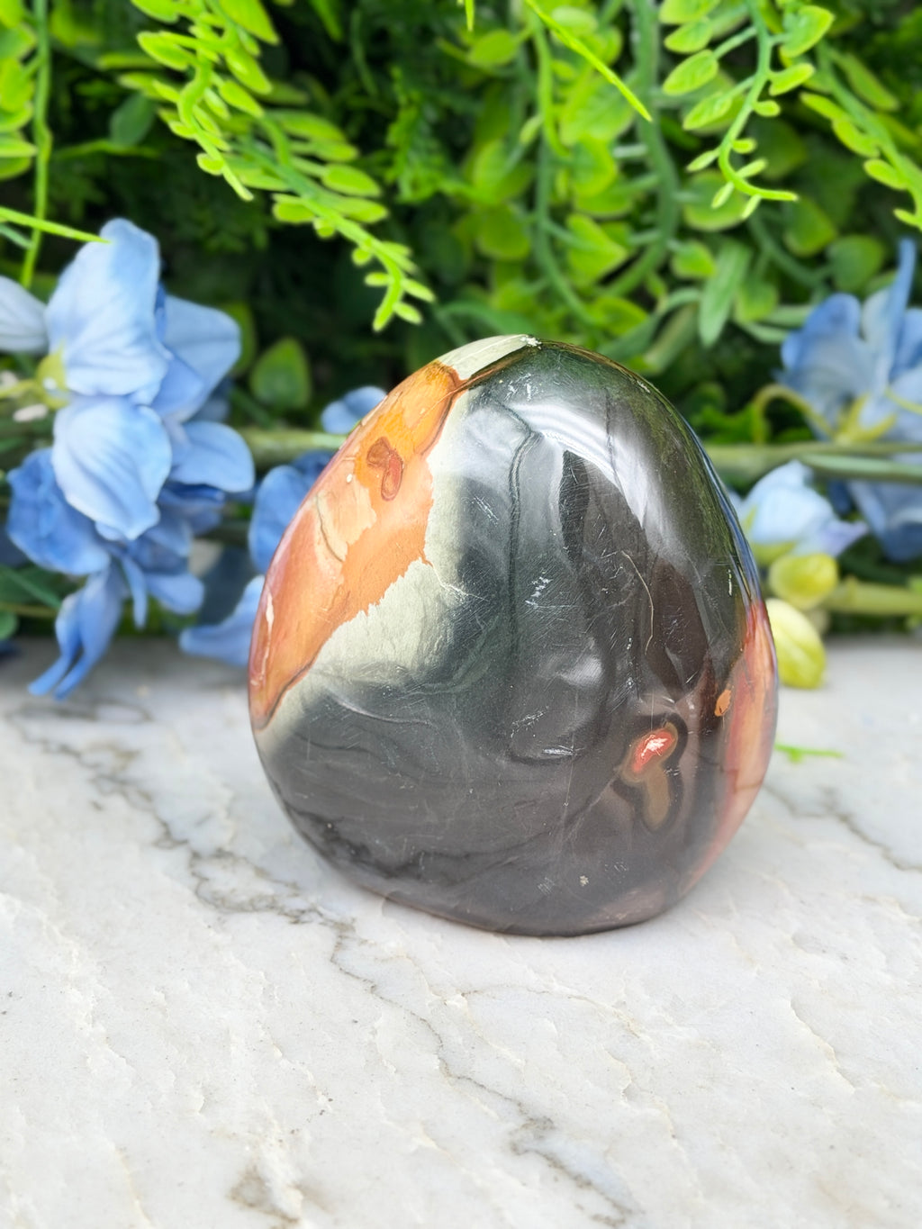 A jadeite gemstone with a black and orange color pattern is displayed on a white marble surface, surrounded by blue flowers.
